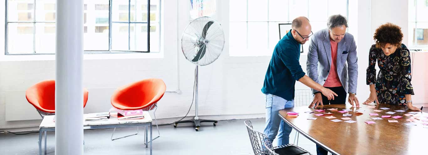 Se muestra un grupo de 3 personas entablando una reunión sobre una mesa de madera Se muestra un grupo de 3 personas entablando una reunión sobre una mesa de madera